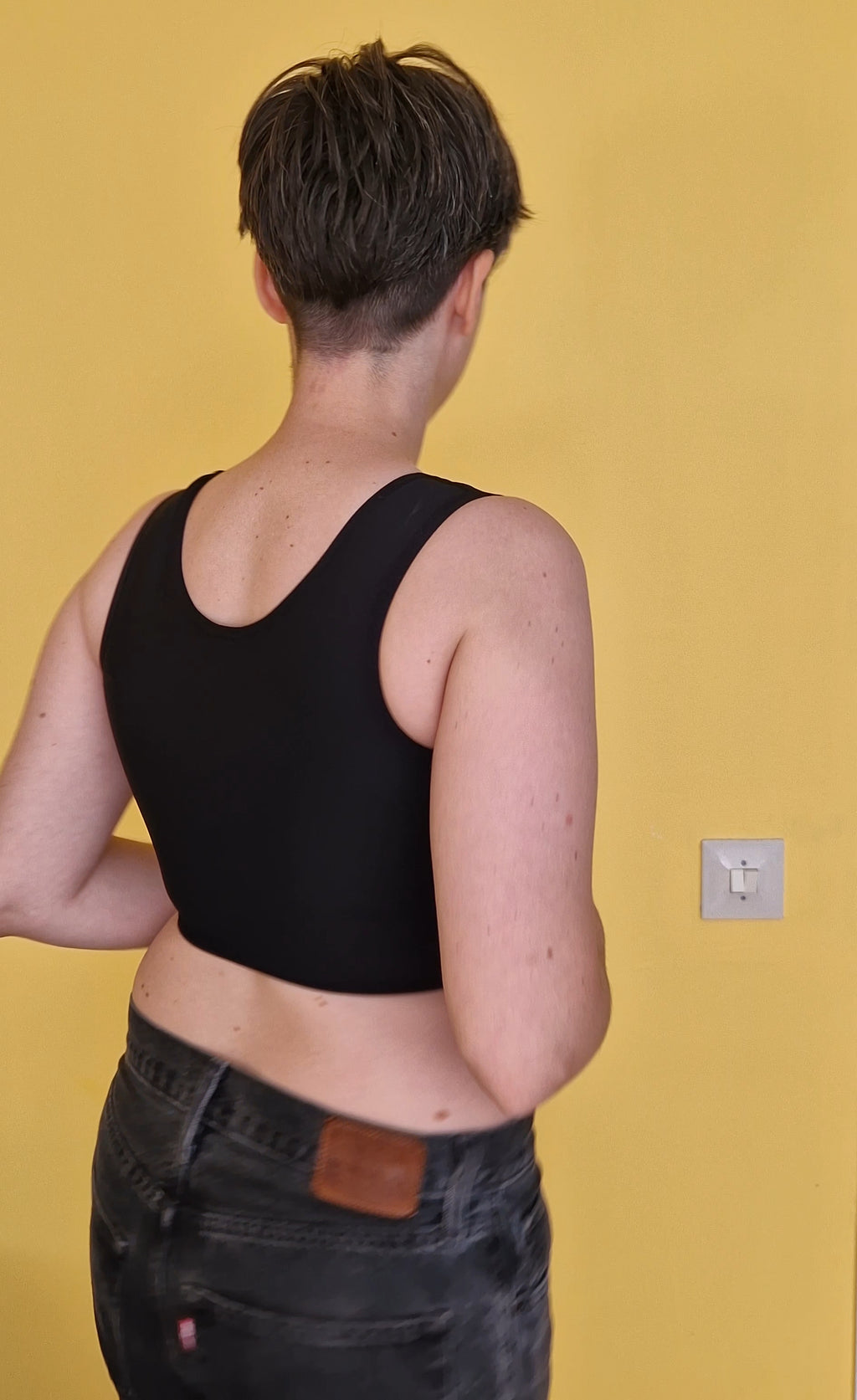 A trans nonbinary person wearing a black short chest compression binder. Back side view.