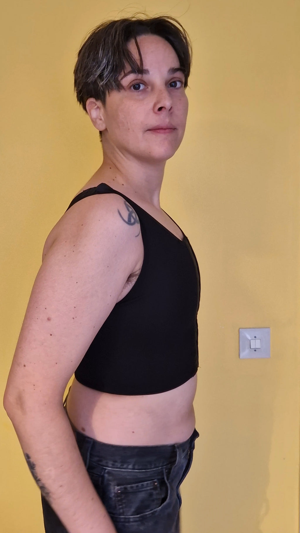 A nonbinary trans person wearing a black post-top-surgery and adjustable binder. Side view