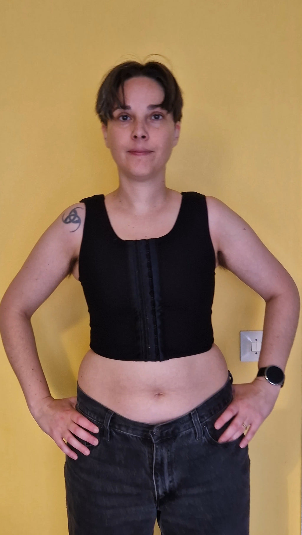 A nonbinary trans person wearing a black post-top-surgery and adjustable binder with clasps on the front and shoulder straps. front view