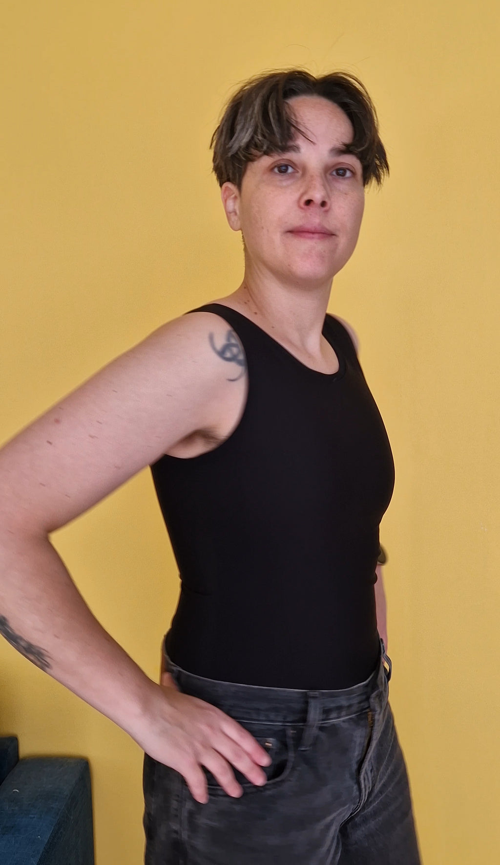 A nonbinary trans person wearing a black long binder that covers the hips, like a tank top. View from the side.