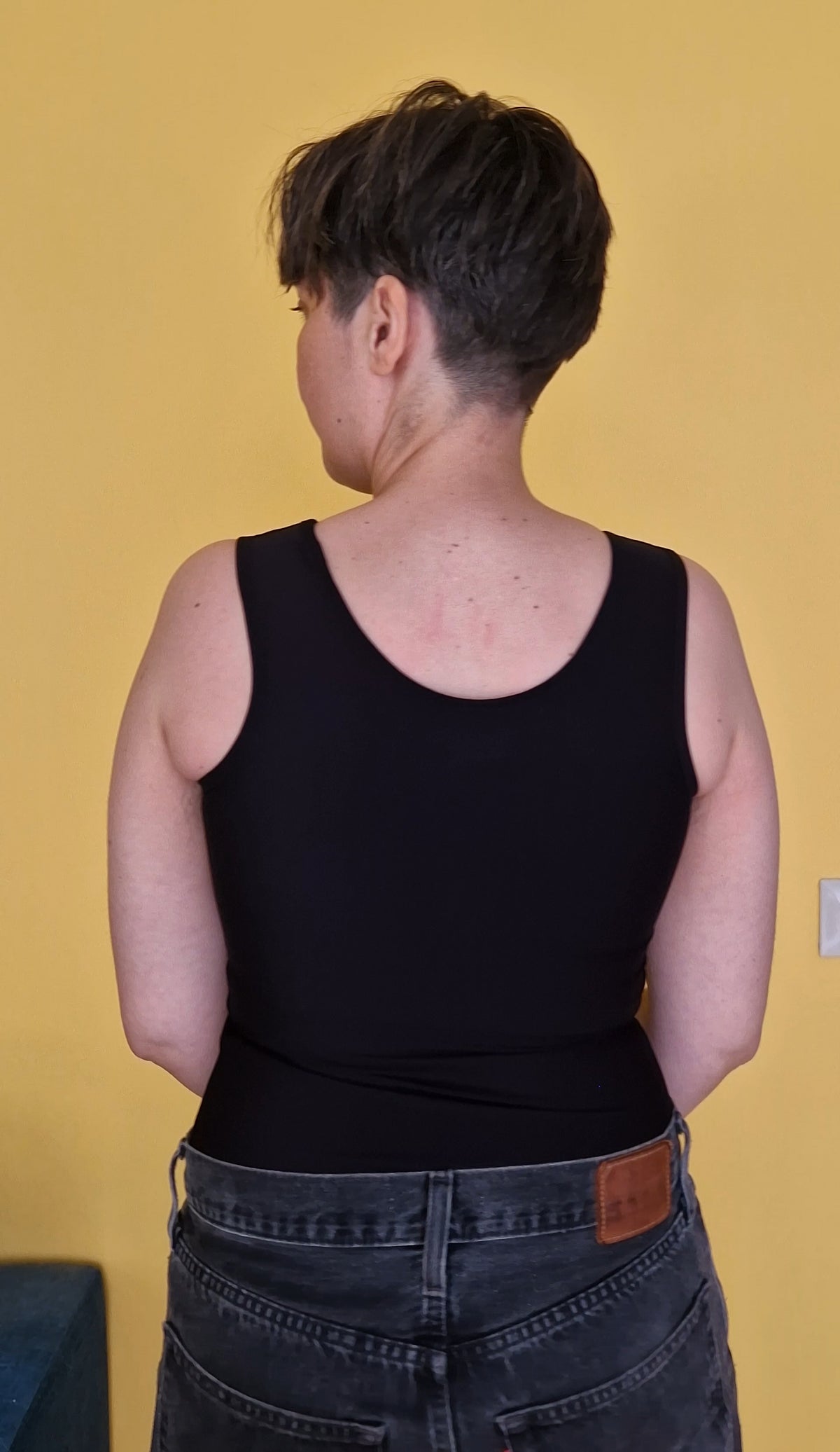 A nonbinary trans person wearing a black long chest binder that covers the hips, like a tank top. View from the back.