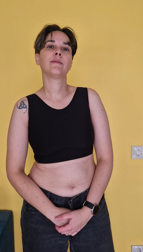 A trans nonbinary person wearing UNTAG sport safe black short chest compression binder. Front view.
