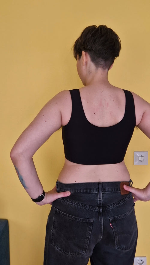 A trans nonbinary person wearing UNTAG sport safe black short chest compression binder. The back view shows a low cut leaving the upper back skin exposed.